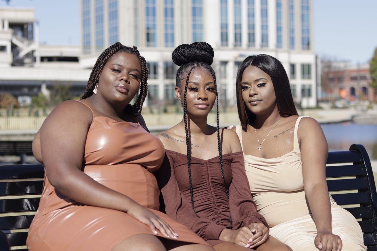 Three African American women in stylish dresses sitting outdoors and posing confidently.